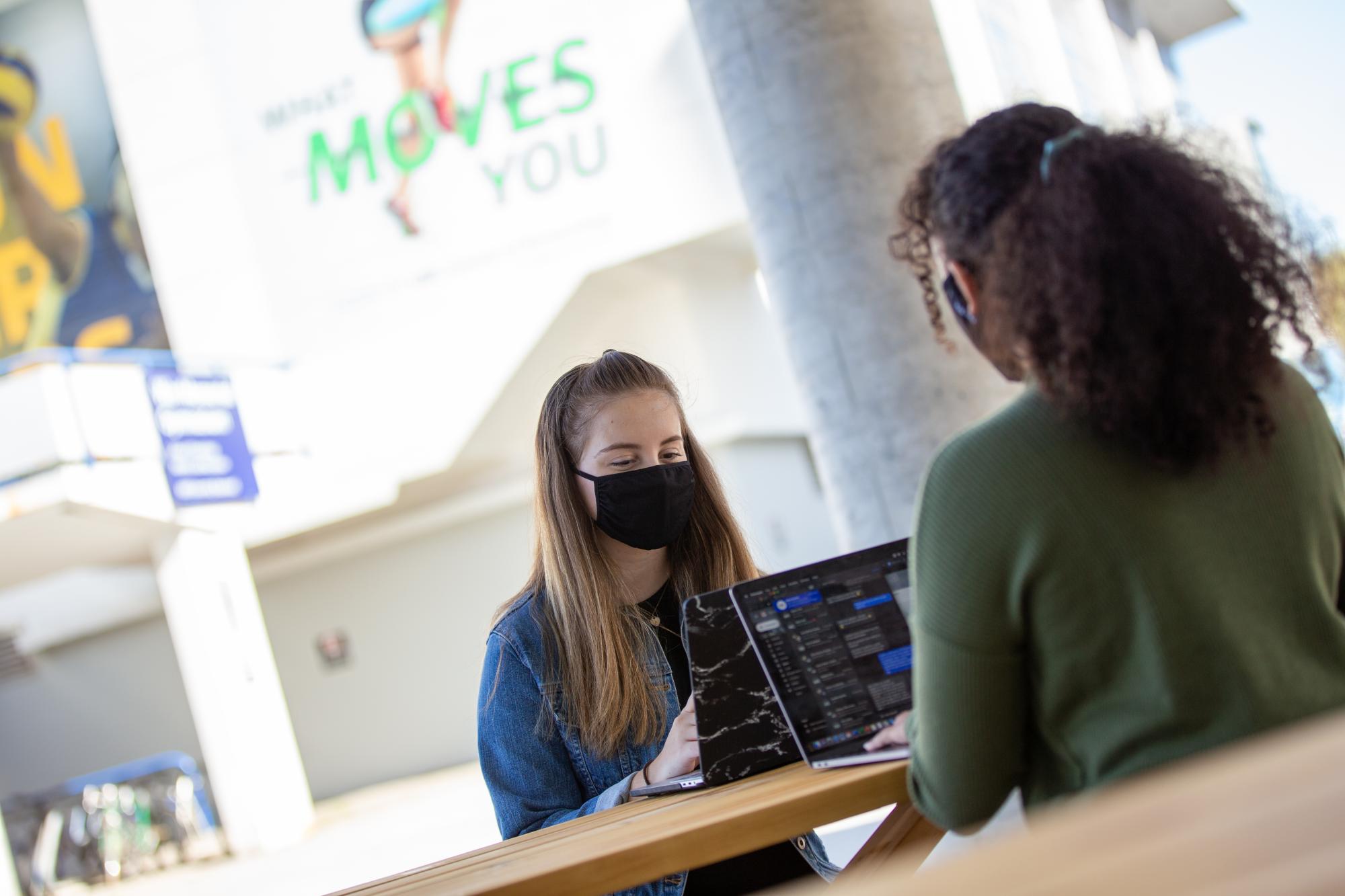 Two students, each wearing a cloth face mask and working on a laptop, sit facing each other across a picnic bench.