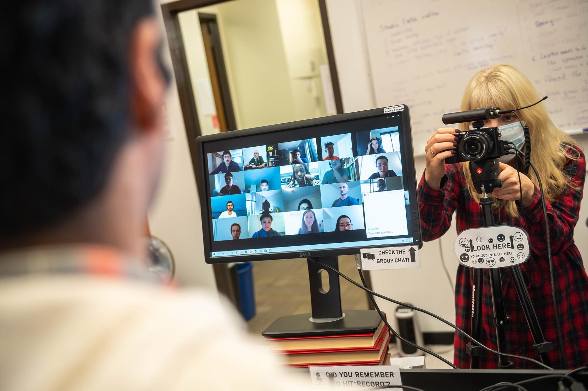 An instructor's point-of-view photo of a computer screen showing a zoom call with many students, while behind the screen is a masked person setting up a video camera facing the instructor. 