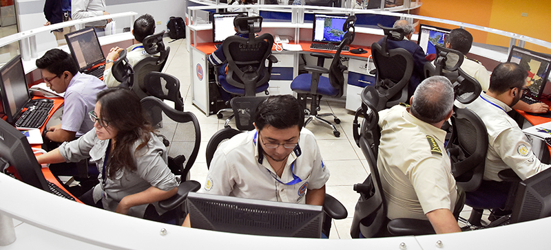 Nine people sit at computer terminals at a round security desk