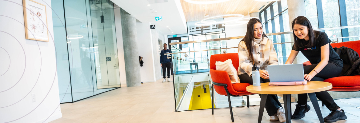 Two students sit in the foreground, looking at a laptop. Another student is walking towards the camera from the rear of the photograph