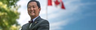 Dr. Santa Ono poses in the foreground, while the Canadian flag flies in the background.
